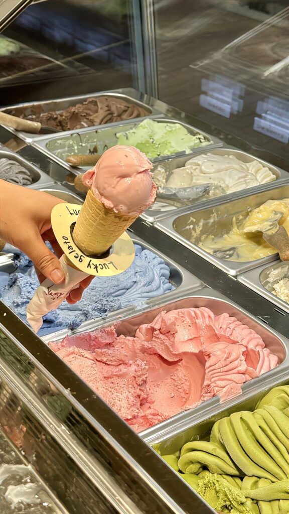 Hand holding pink gelato cone over display of colorful ice cream flavors in shop.