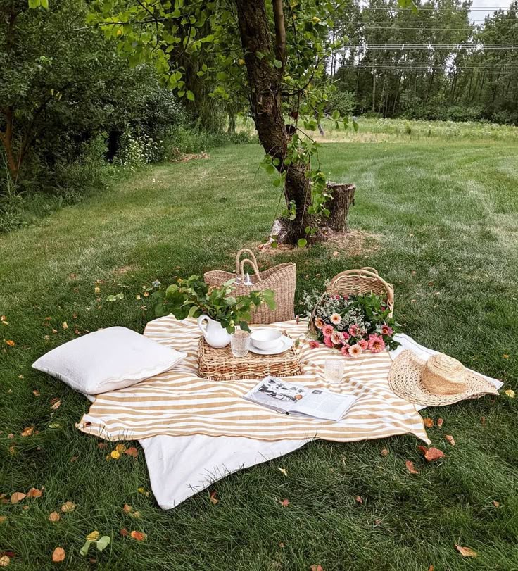 Cozy picnic setup under a tree with a striped blanket, flowers, wicker baskets, tea set, and a straw hat on lush green grass.