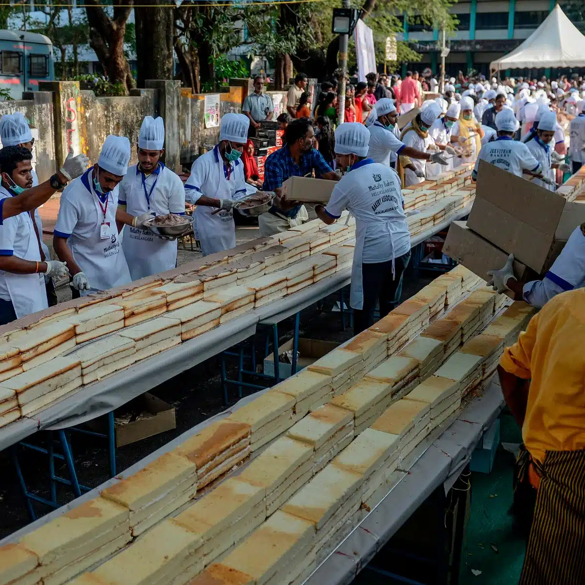 Chefs in uniforms assembling a large cake outdoors at a public event, demonstrating teamwork and baking skills.