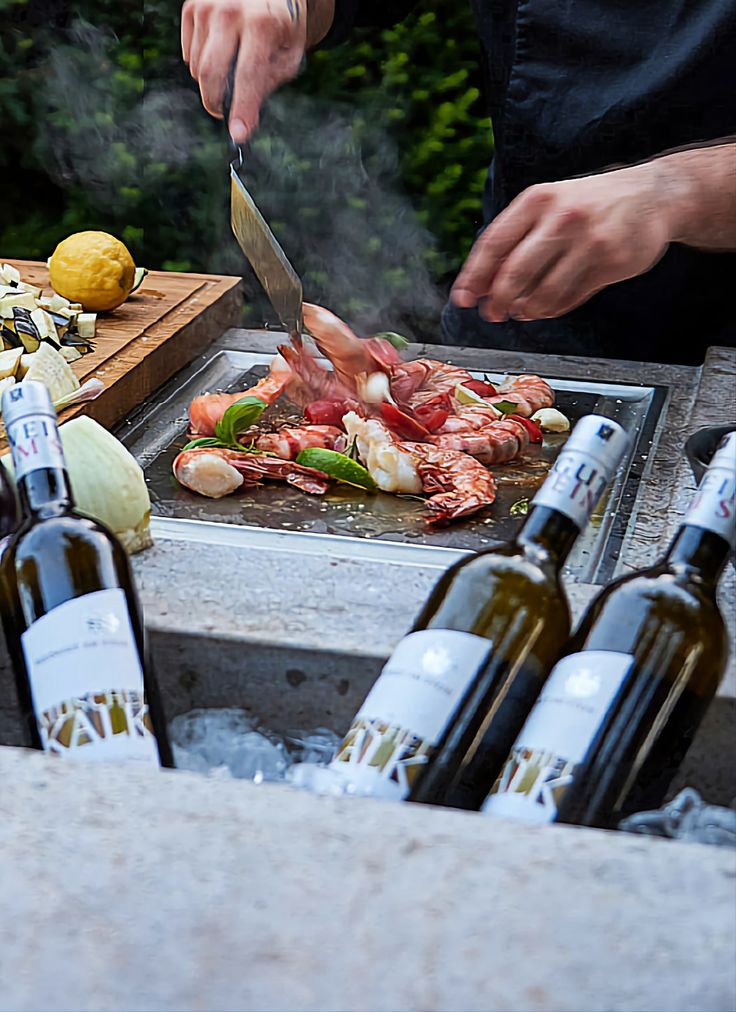 Chef grilling seafood on outdoor cooktop near wine bottles, with steam rising and fresh ingredients nearby.