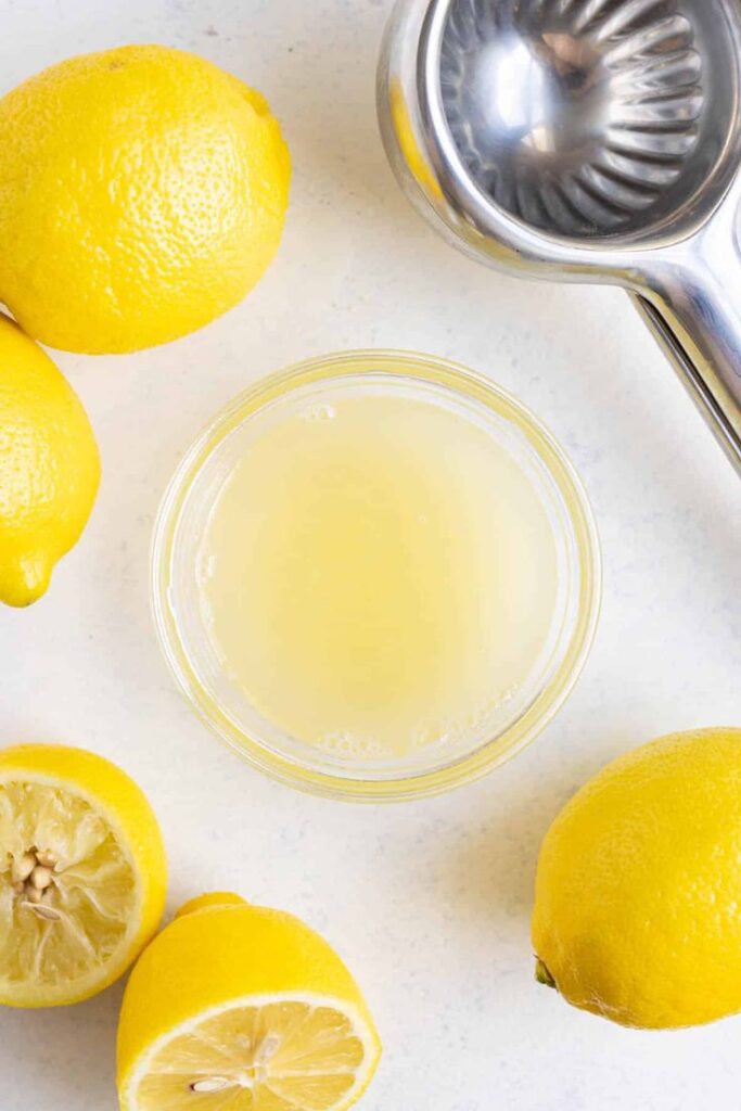 Fresh lemon juice in a bowl surrounded by whole and halved lemons with a metal juicer on a countertop.
