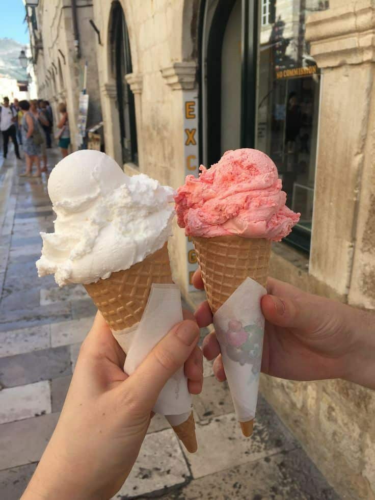 Two people holding vanilla and strawberry ice cream cones against a historic cobblestone street background.