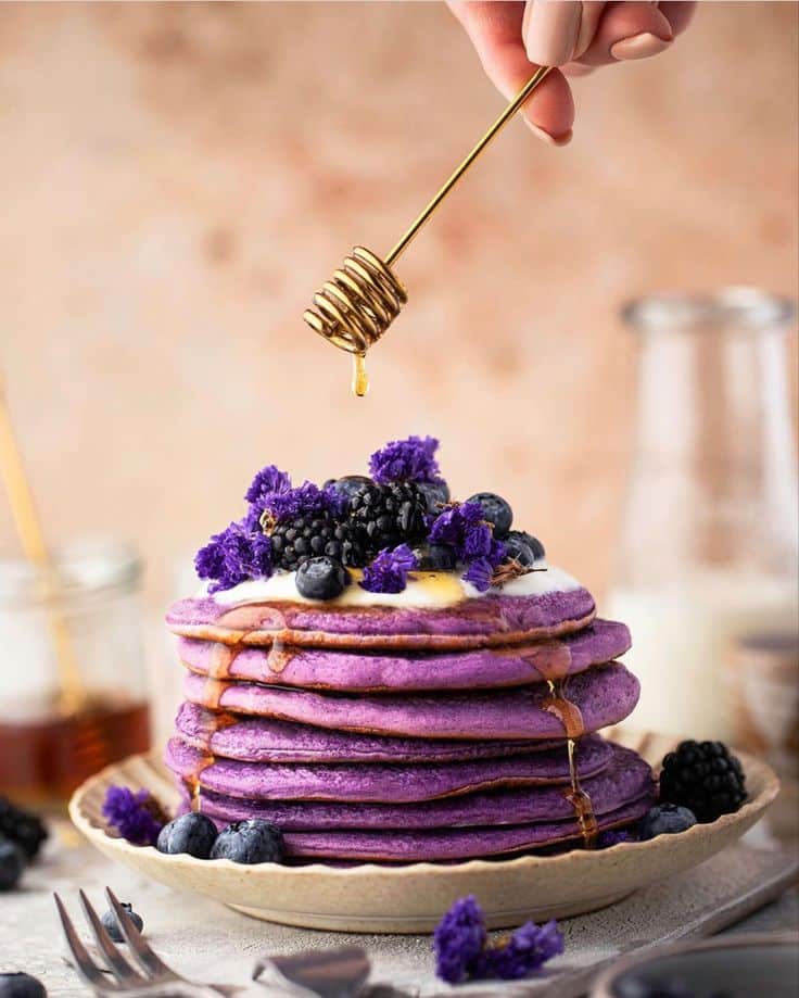 Vibrant purple pancakes with honey drizzle, topped with berries and flowers on a rustic plate. Perfect breakfast treat!