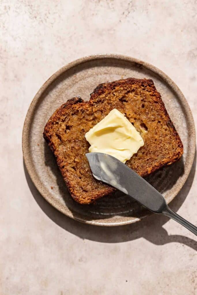 Banana bread slice with butter on a ceramic plate with knife, perfect for breakfast or snack.