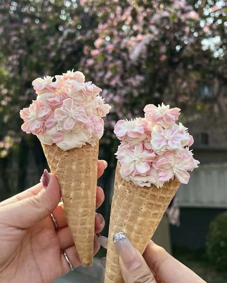Two waffle cones topped with floral pink and white ice cream against a blurred tree background.