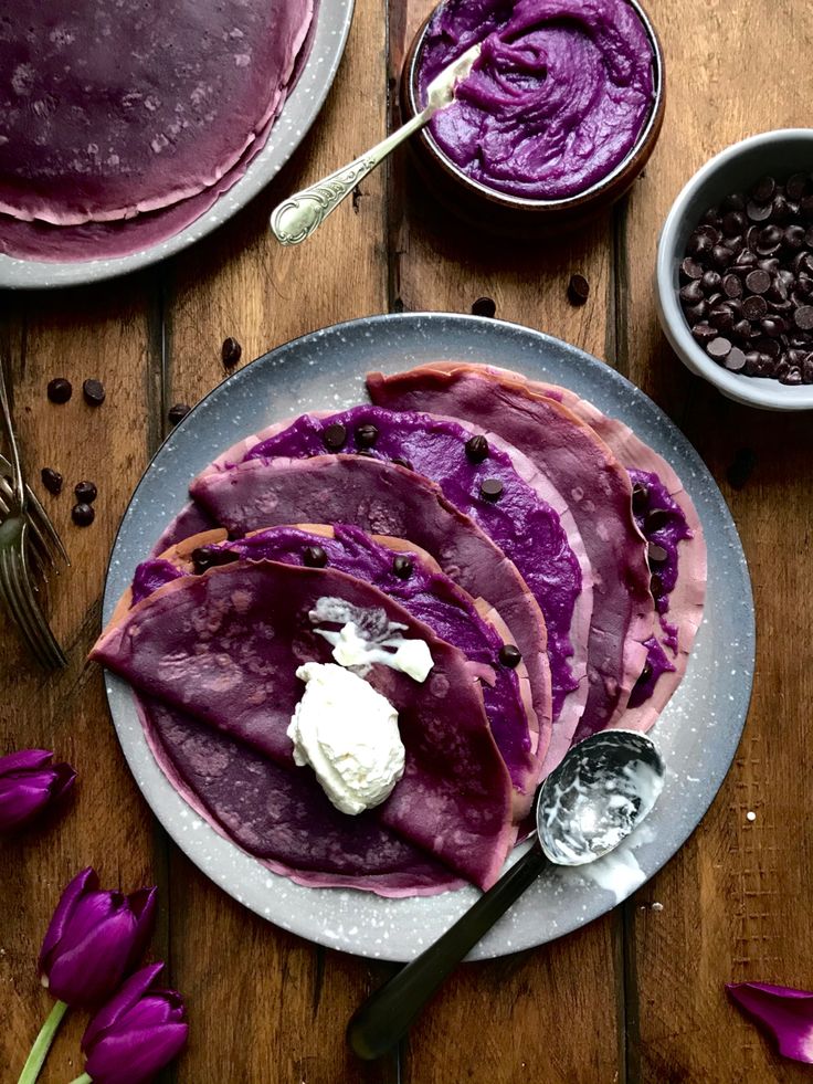Purple crepes with whipped cream on a plate, surrounded by chocolate chips and flowers on a wooden table.