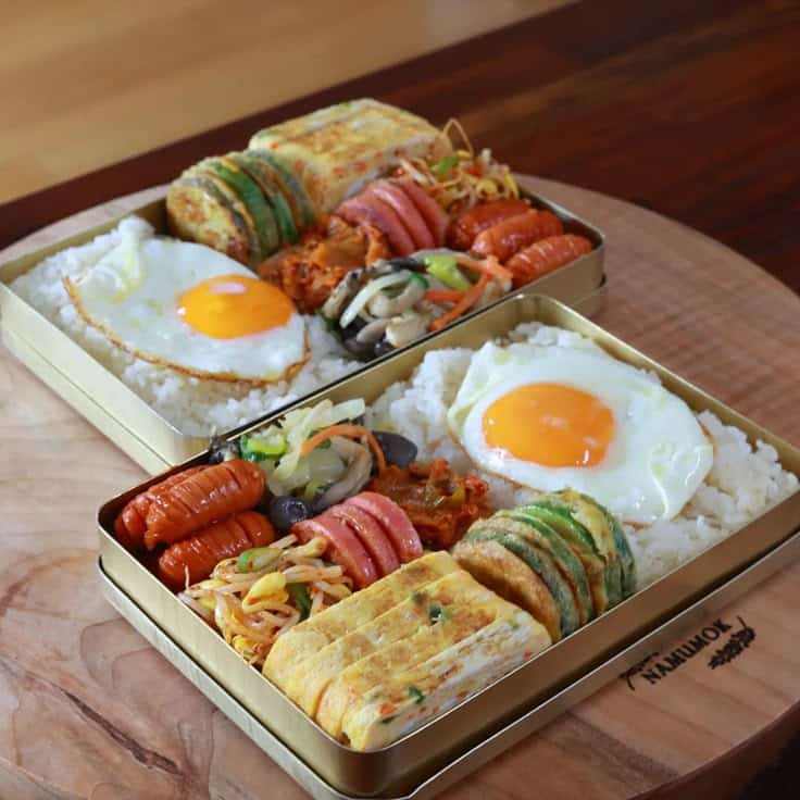 Bento box with fried eggs, rice, vegetables, and sausage arranged on a wooden surface, showcasing a colorful meal.