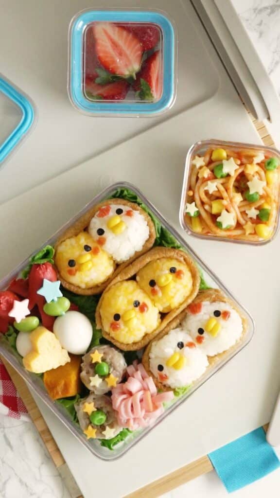Cute bento box with rice shaped like chicks, colorful vegetables, pasta, and strawberry dessert on a tray.
