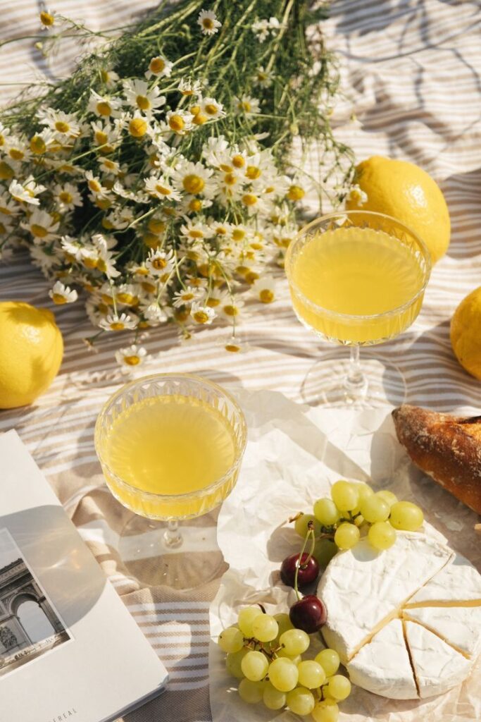 Sunny picnic setup with yellow drinks, Brie cheese, grapes, wildflowers, and lemons on a striped cloth.