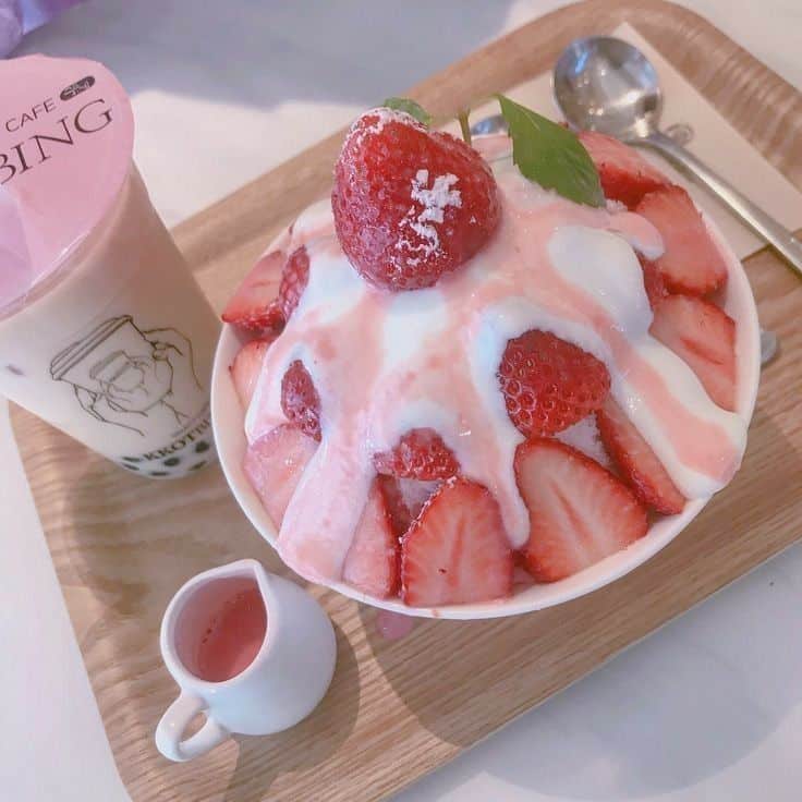 Strawberry bingsu with fresh strawberries, cream, and strawberry sauce, served with a drink on a wooden tray.
