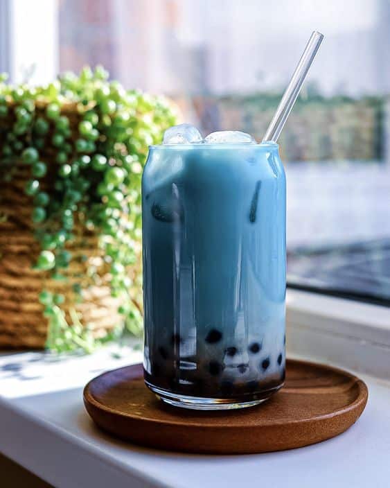 Blue bubble milk tea with ice and tapioca pearls in a glass jar, beside a plant on a sunlit windowsill.