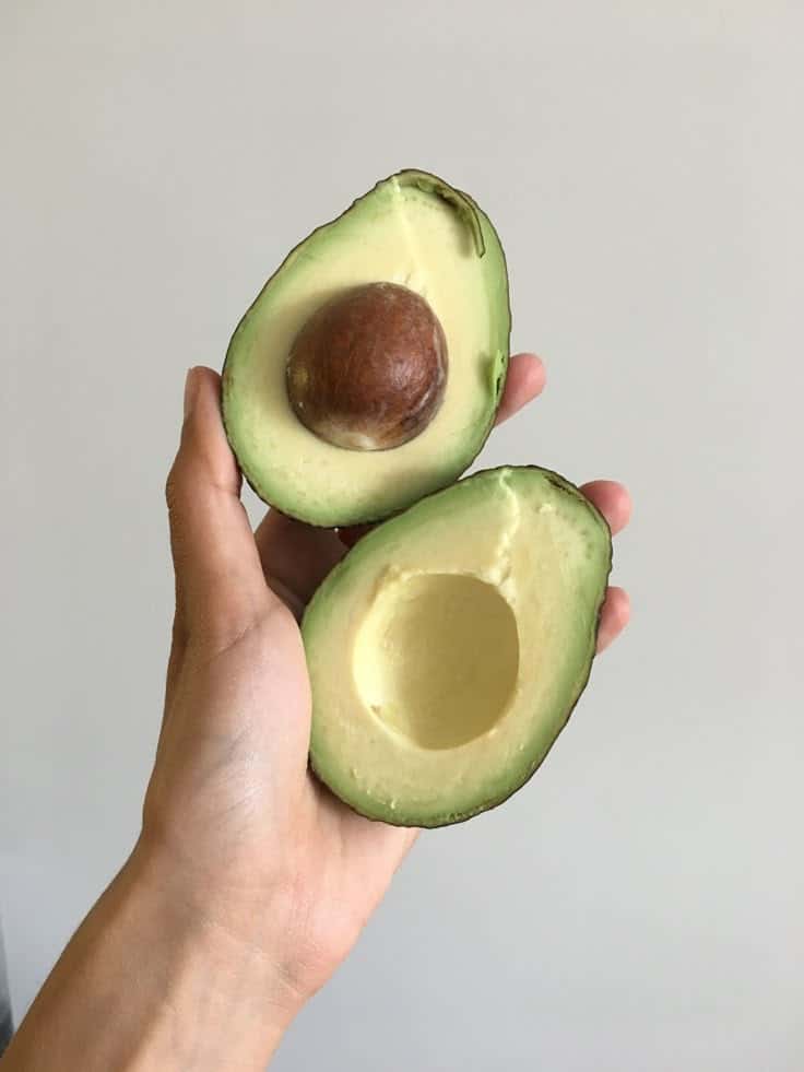Hand holding a fresh avocado cut in half with seed visible against a plain background.
