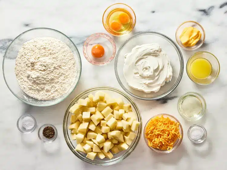 Bowls of flour, eggs, sour cream, chopped potatoes, cheese, and seasonings on a marble countertop.