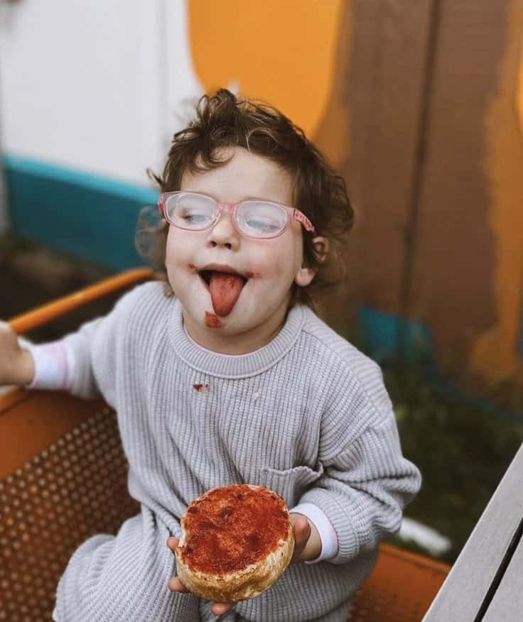 Child with glasses enjoying a jam-covered bun, smiling with a playful expression outdoors.