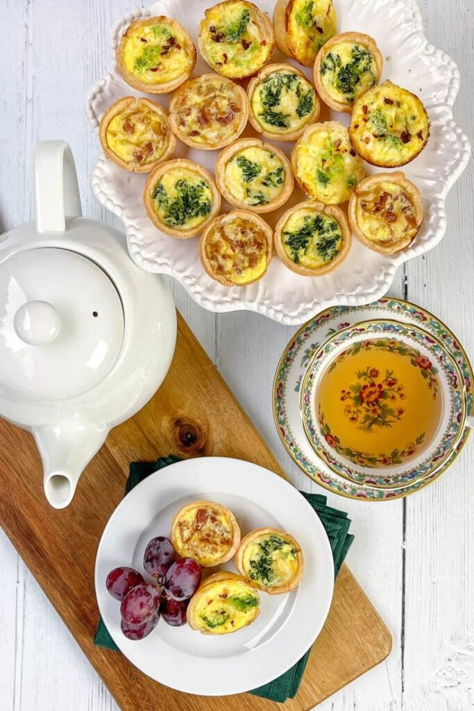 Mini quiches on a plate with a teapot and a cup of tea, garnished with grapes, on a wooden board.