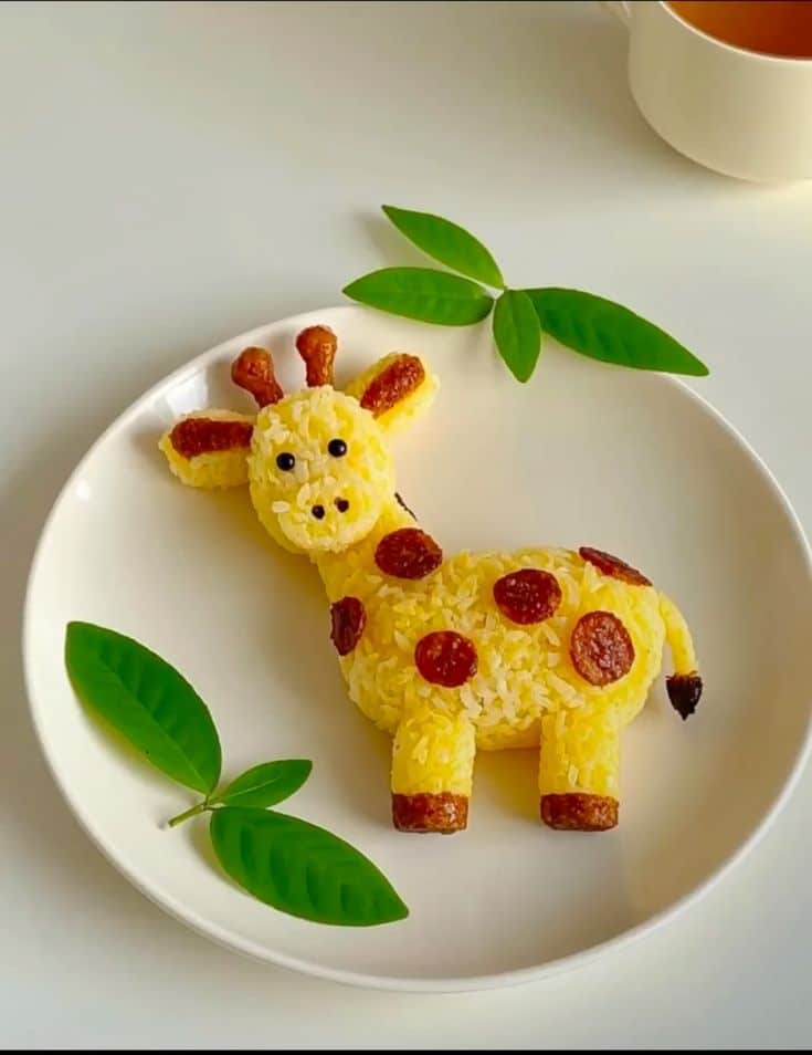 Creative giraffe-shaped rice dish with sausage spots and garnish leaves on a white plate.