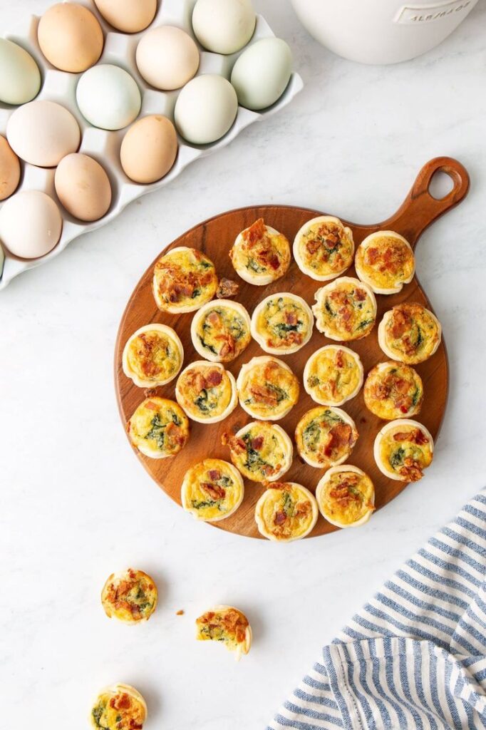 Mini quiches on a wooden board with a striped cloth and fresh eggs in the background. Perfect for brunch.