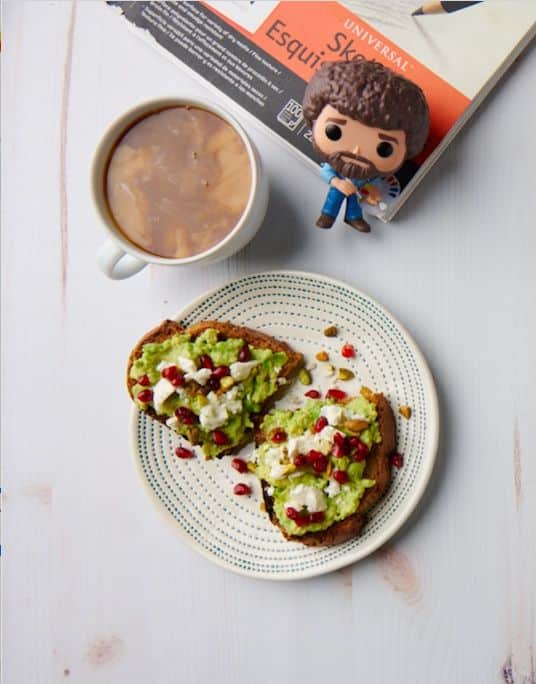 Screenshot - Wake Up Right: 10 Easy Toast Recipes Packed with Energy | Avocado toast with pomegranate and feta on a plate next to coffee and a small figurine on a white table.