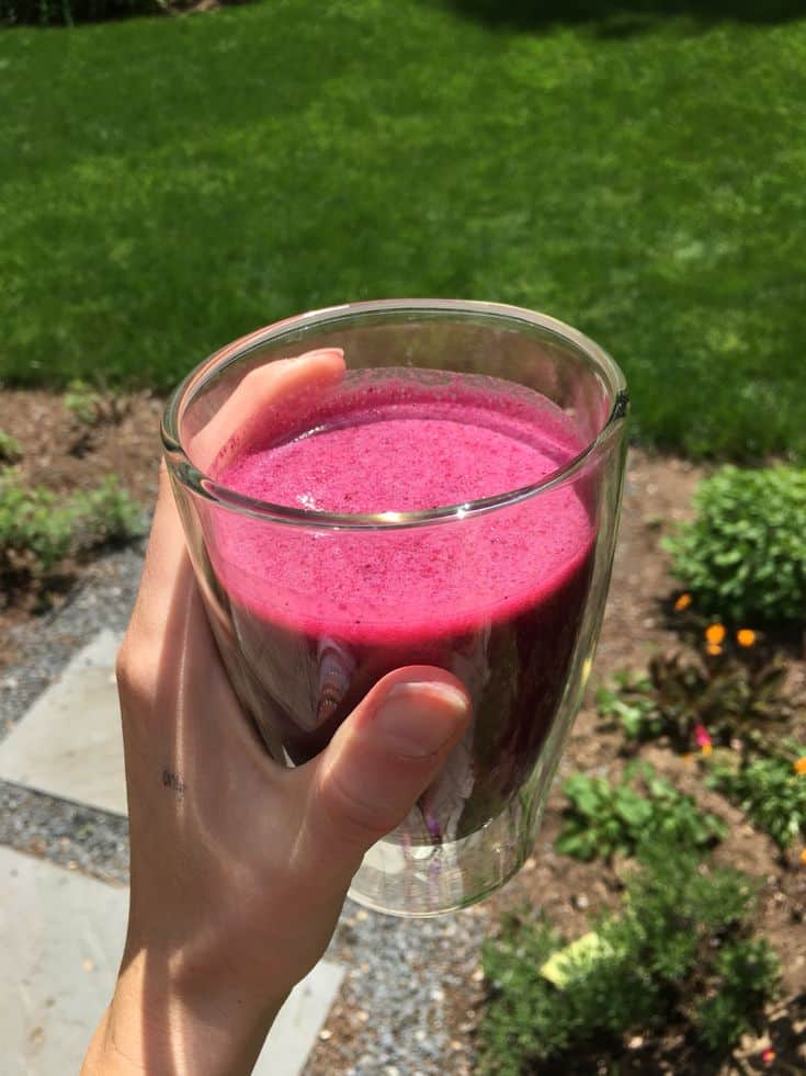 Hand holding a glass of bright pink smoothie in a sunny garden setting.