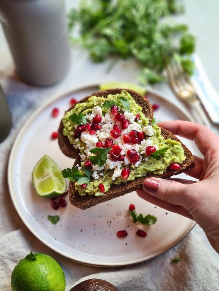 Ben jij op zoek naar een lekker lunchrecept_ Deze - Wake Up Right: 10 Easy Toast Recipes Packed with Energy | Avocado toast topped with feta, pomegranate, and cilantro on a plate with lime, held by a hand for a fresh breakfast.