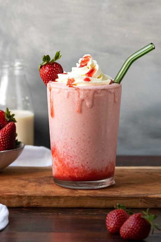 Strawberry milkshake with whipped cream and fresh strawberries, served with a straw on a rustic wooden board.