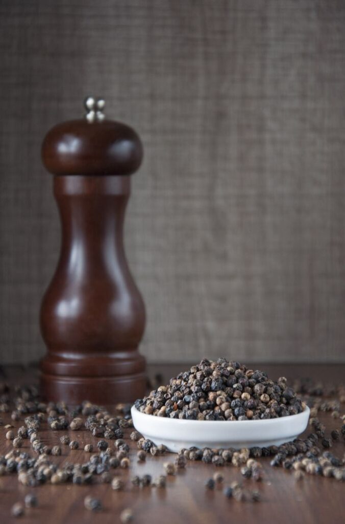 Bowl of whole black peppercorns next to a wooden pepper grinder on a table.
