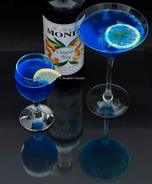 Blue Curacao cocktails in glasses with lemon slices, bottle in background, reflecting on glass surface.