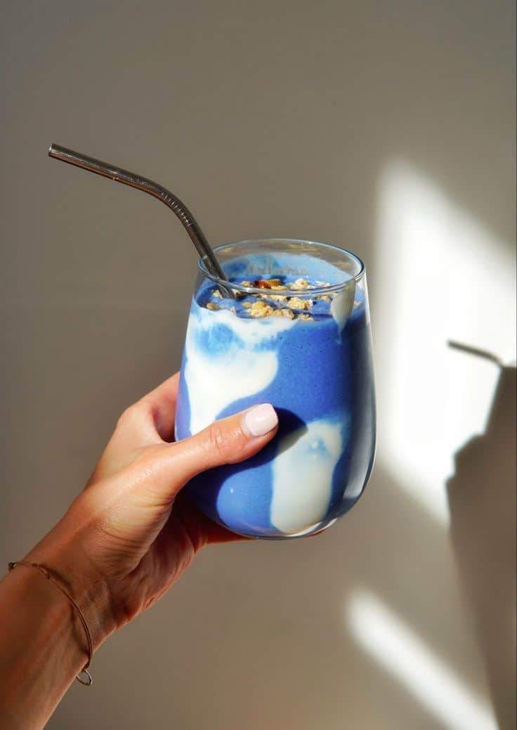 Hand holding a vibrant blue smoothie with a metal straw, garnished with granola, in natural sunlight.