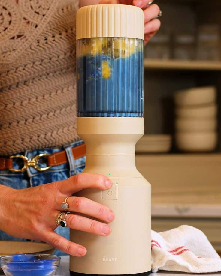 Person blending blue smoothie in a beige blender in a kitchen setting.