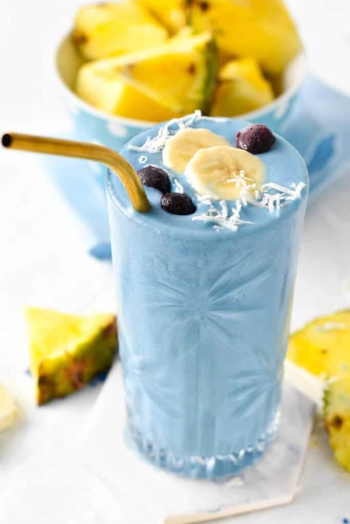 Blue smoothie topped with banana slices, berries, and coconut flakes, paired with fresh pineapple in the background.