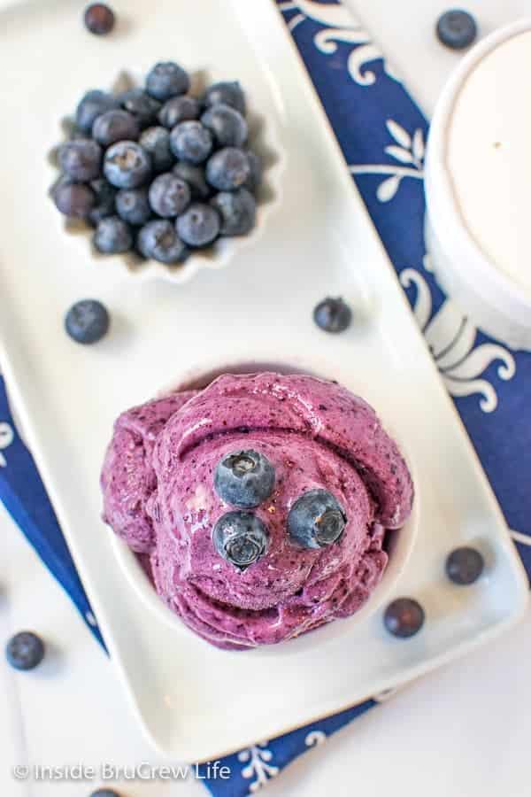 Blueberry ice cream in a bowl topped with fresh blueberries on a white tray, next to a dish of blueberries.