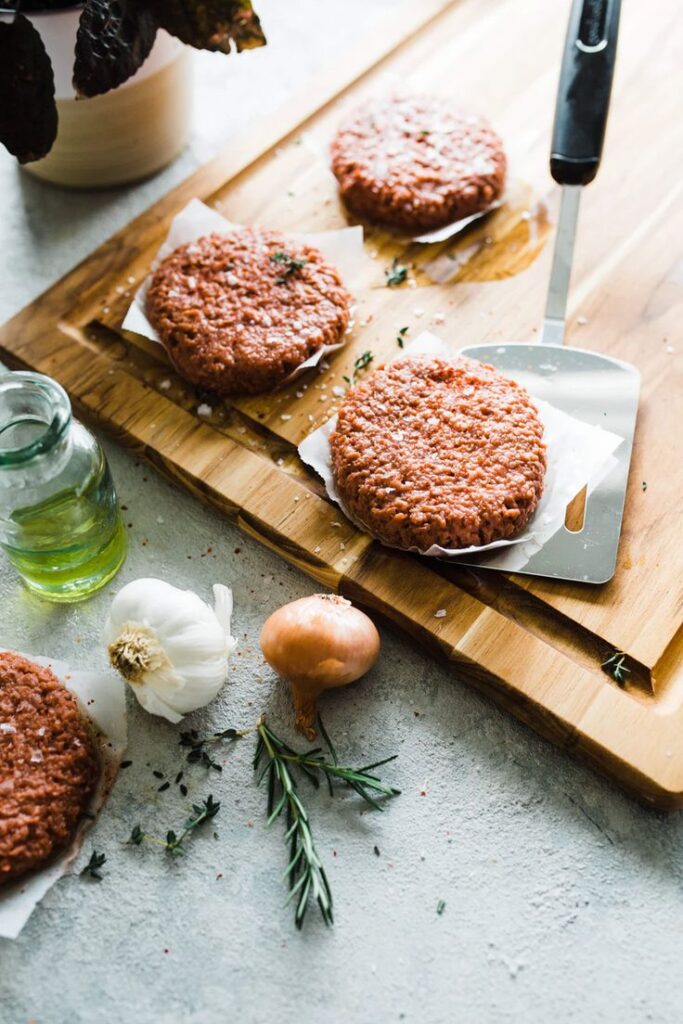 Raw burger patties on a wooden board with garlic, onion, and herbs, ready for cooking preparation.