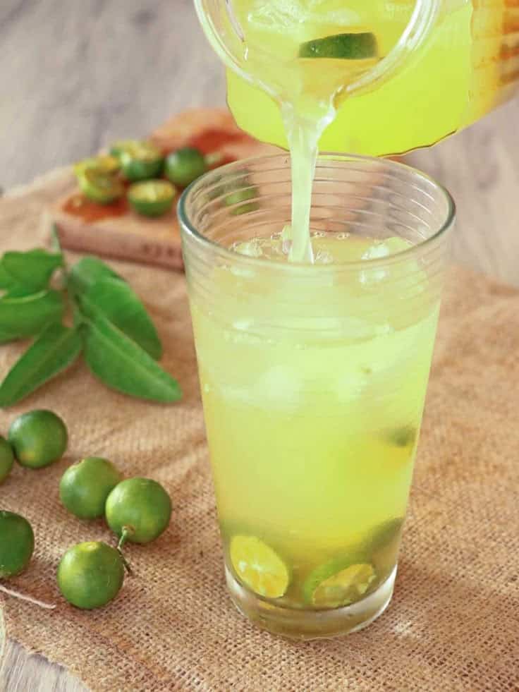 Refreshing calamansi juice poured into a glass with ice cubes, surrounded by fresh calamansi fruits on burlap.