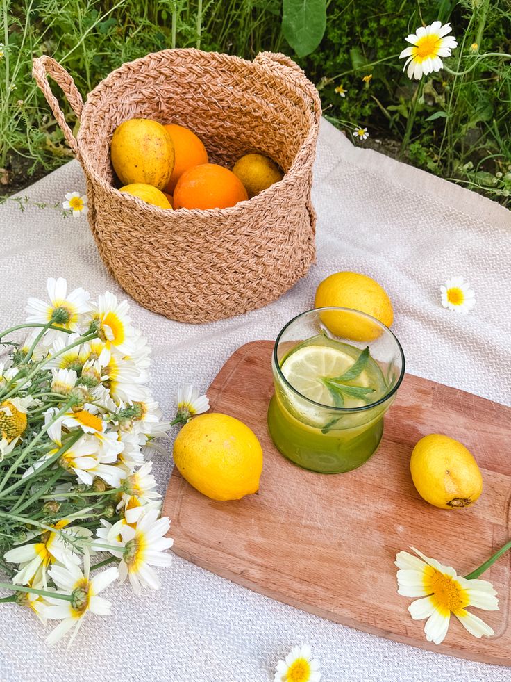 Basket of citrus fruits with lemons, daisies, and green juice on a picnic setup outdoors.
