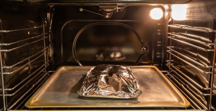 Foil-wrapped food baking on a tray in a lit oven for cooking or roasting.