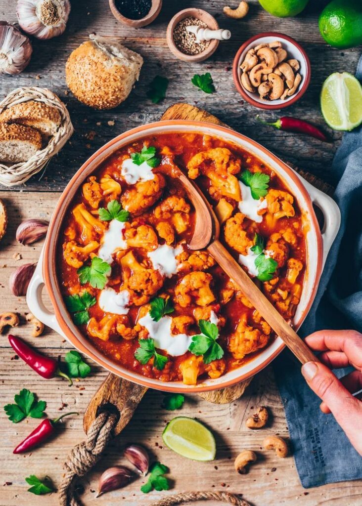 Vegetable curry with cauliflower in a bowl, garnished with herbs, surrounded by bread and spices on a wooden table.