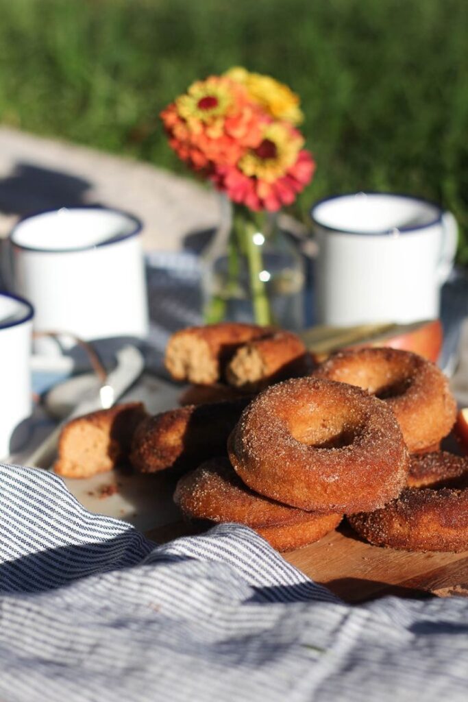 Cinnamon sugar donuts with enamel mugs and flowers on wooden table in a sunny outdoor setting.