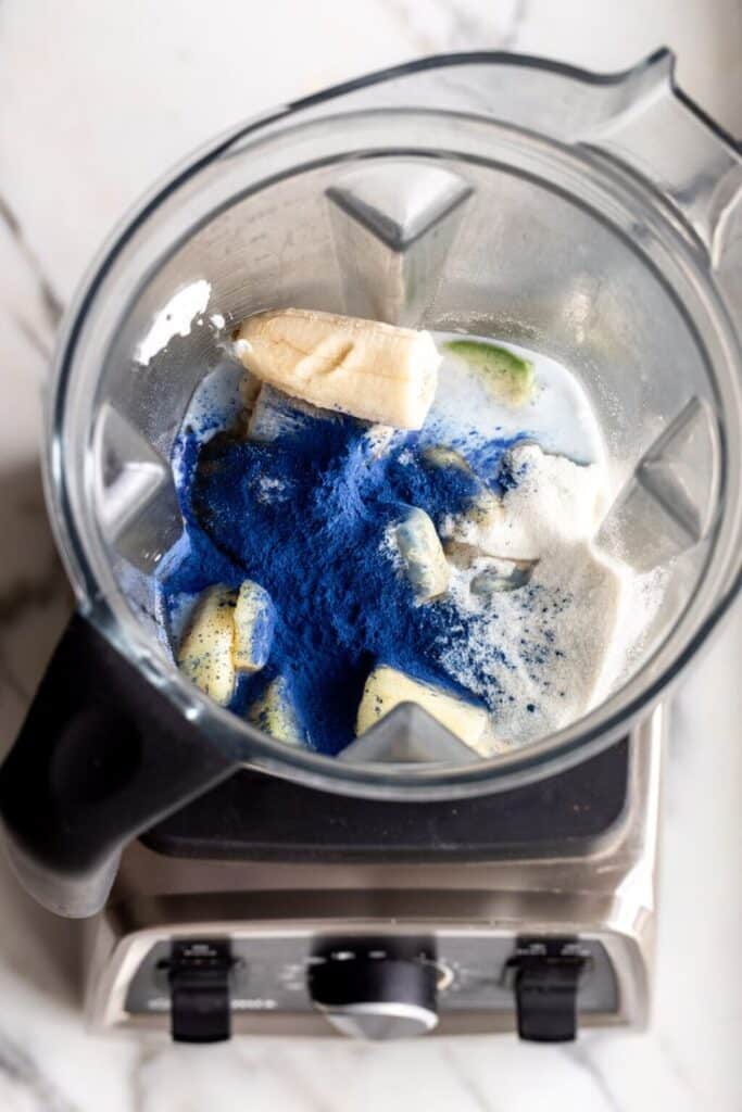 Blender with banana, blue powder, and milk blend ingredients on countertop.