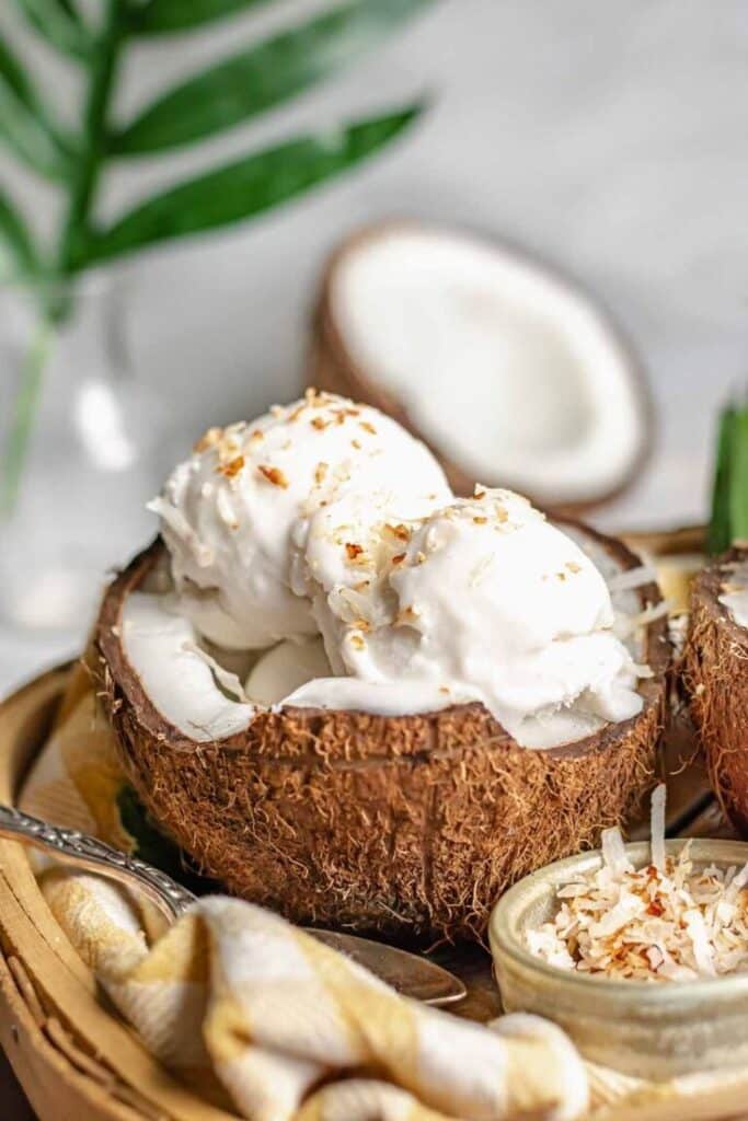 Coconut ice cream served in a coconut shell with a topping of toasted coconut flakes on a wooden tray.