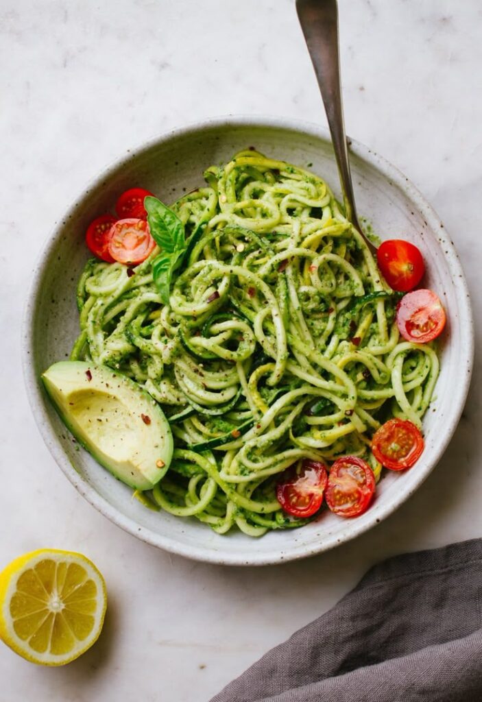 Zucchini noodles with avocado, cherry tomatoes, and basil in a bowl, garnished with lemon for a fresh summer dish.