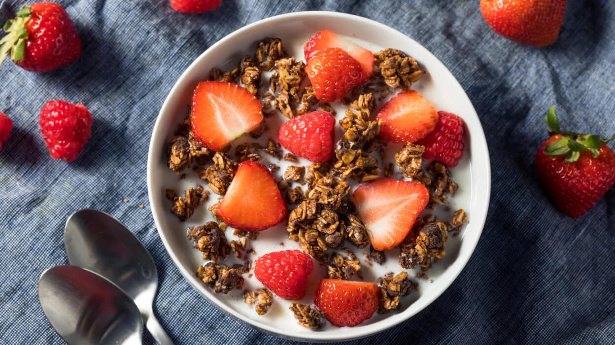 Bowl of granola with fresh strawberries and raspberries on yogurt, surrounded by scattered berries and spoons.