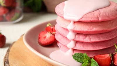 Stack of pink pancakes drizzled with sauce, garnished with strawberries and mint on a plate.
