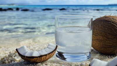 Glass of coconut water on sandy beach with coconut halves near the ocean.