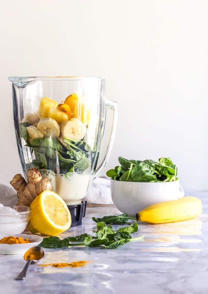 Blender with fresh ingredients for a healthy green smoothie, including spinach, banana, ginger, and lemon on a countertop.