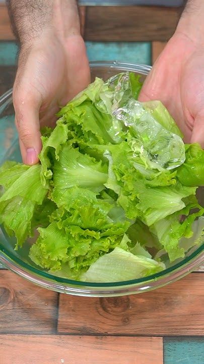 Hands washing fresh green lettuce leaves in a glass bowl with water on a wooden table.