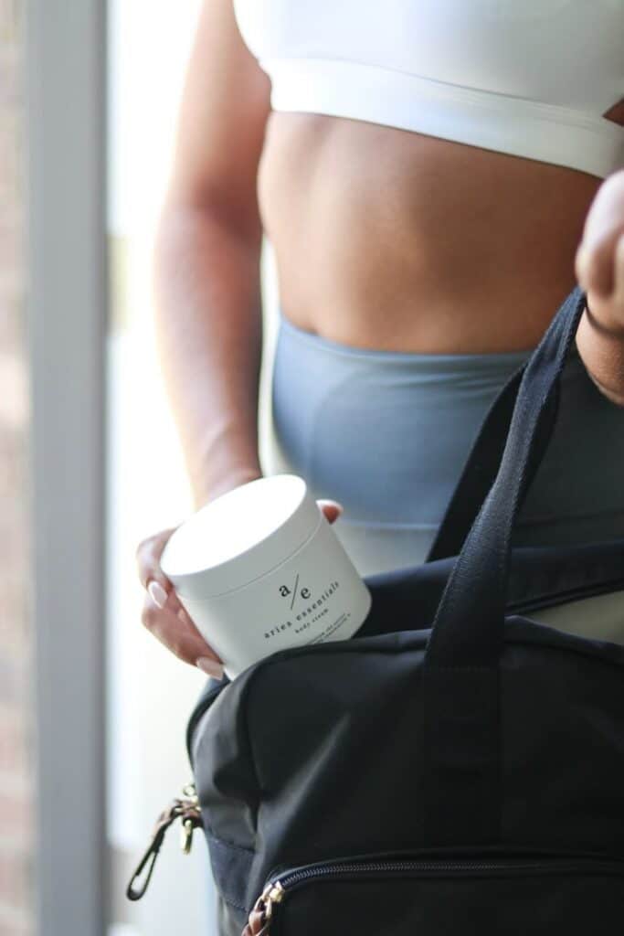 Woman in sportswear placing Aries Essentials body scrub into gym bag.