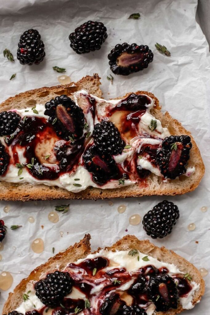 Rustic bread topped with cream cheese, fresh blackberries, and thyme, drizzled with honey on parchment paper.