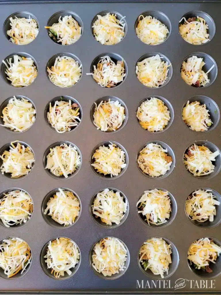 Mini muffin pan filled with shredded cheese-topped appetizers ready for baking. Perfect for parties or snacks.