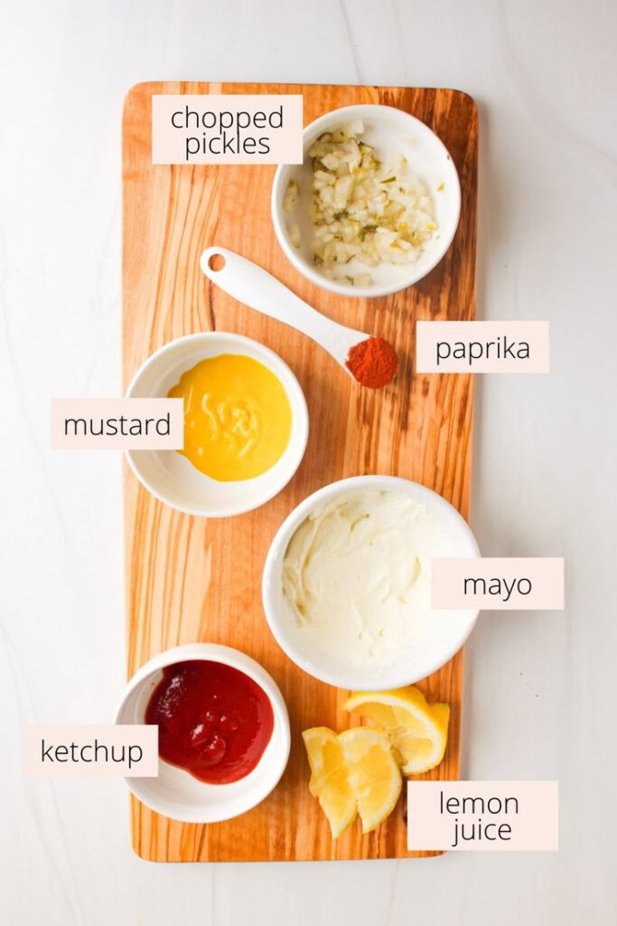 Ingredients for homemade sauce on a wooden board: chopped pickles, mayo, mustard, ketchup, paprika, and lemon wedges.