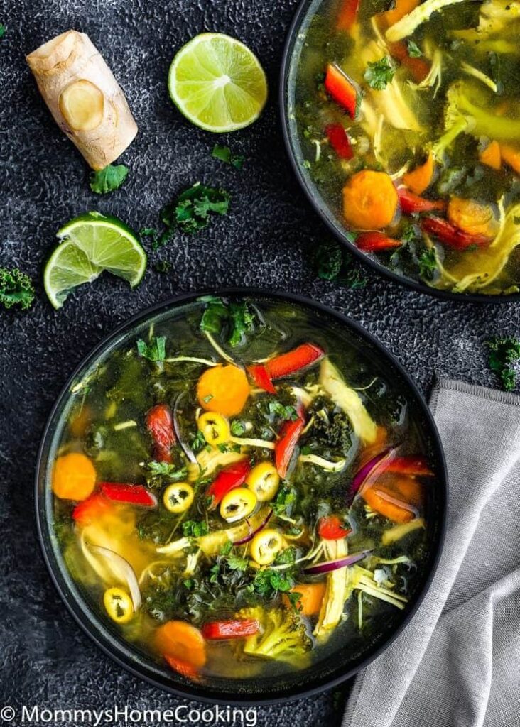 Two bowls of colorful vegetable soup with lime, ginger, and herbs on a dark textured background.